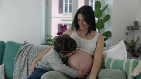 Foto de Niño besando el vientre embarazada de la madre sentado en el sofá en casa en conmovedor gesto genuino de amor y afecto hacia el hermano bebé nonato durante la etapa final del embarazo - Imagen libre de derechos