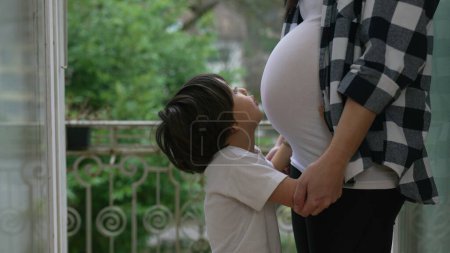 Kleiner Junge drückt Liebe aus, indem er den Schwangerschaftsbauch der Mutter küsst, zärtlicher Moment im dritten Trimester auf dem heimischen Balkon, Bruder umarmt ungeborenes Baby
