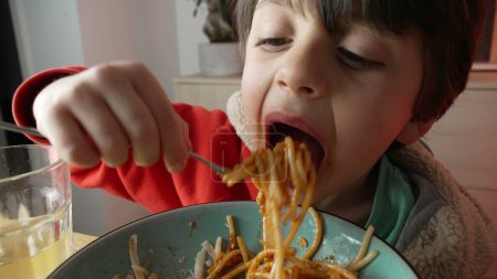 Child grabs fork and eats a mouthful of pasta spaghetti for supper, macro close-up face in wide angle, Italian food