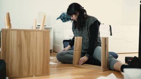 Téléchargez les photos : Table de chevet en bois de montage de jeune femme, démontrant l'expertise de Do-It-Yourself dans le décor à la maison, parfait pour les nouveaux propriétaires commençant frais - rénovation domiciliaire - en image libre de droit