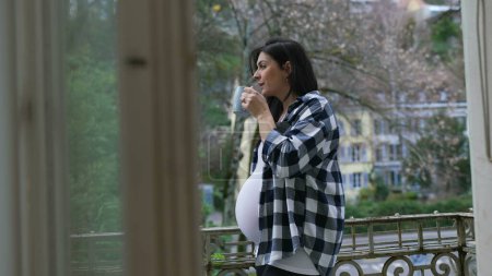 Photo for Contemplative pregnant woman stands by apartment balcony sipping tea looking at view from residence, enjoying quiet moment during third trimester - Royalty Free Image