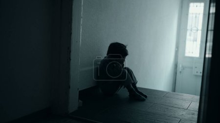 Foto de Niño de cinco años que lidia con la desesperación emocional, sentado solo en el corredor oscuro, cubriendo la cara con las manos, la depresión infantil conceptualizada - Imagen libre de derechos