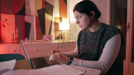 Téléchargez les photos : Femme travaillant sur ordinateur portable à la maison le soir, 30s personne s'engageant dans le travail ou l'étude à distance, focalisé sur l'écran d'ordinateur la nuit - en image libre de droit
