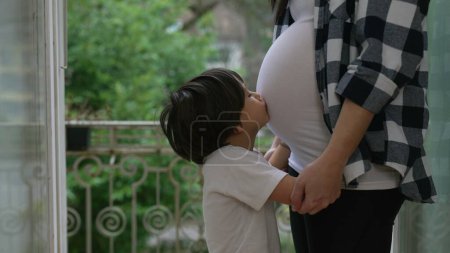 Kleiner Junge drückt Liebe aus, indem er den Schwangerschaftsbauch der Mutter küsst, zärtlicher Moment im dritten Trimester auf dem heimischen Balkon, Bruder umarmt ungeborenes Baby