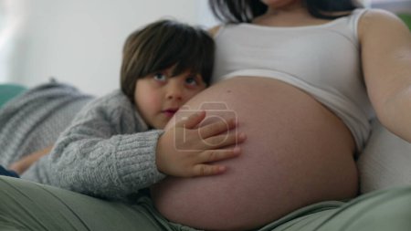 Photo for Child gently rubbing mother's pregnant belly during third trimester of pregnancy, happy heartwarming family moment, close-up face and hand - Royalty Free Image