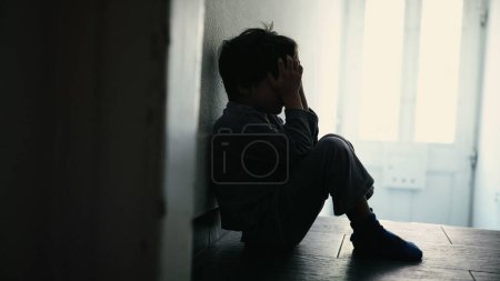 Foto de Pequeño niño cubriendo la cara sintiendo desesperación Un niño pequeño sentado en el pasillo oscuro Corredor en casa sufriendo solo en crisis - Imagen libre de derechos