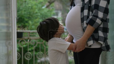 Pequeño niño expresando amor por besar el vientre embarazada de la madre, tierno momento del tercer trimestre en el balcón casero, hermano abrazando al bebé nonato