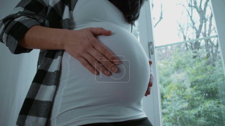 Foto de Escena de embarazo dichosa, mujer alegre embarazada tocando suavemente su vientre embarazada de 8 meses, soñando con el recién nacido cerca de una ventana soleada - Imagen libre de derechos
