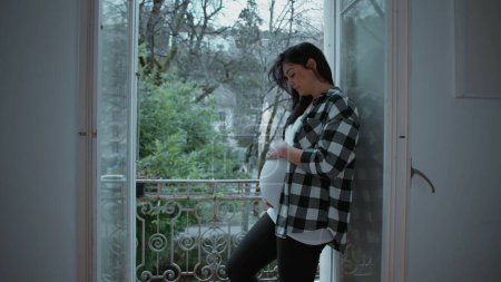 Téléchargez les photos : Mère enceinte caressant tendrement son ventre de 8 mois par appartement balcon, rayonnant d'amour et d'affection tout en attendant le nouveau-né - en image libre de droit