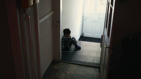 Téléchargez les photos : Petit garçon en crise, assis seul dans le couloir ombragé de la maison, se cacher le visage dans les mains, vivre un profond désespoir et l'isolement, enfant aux prises avec la honte seule, concept de dépression infantile - en image libre de droit