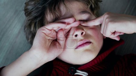 Foto de Retrato de cerca del niño fatigado tocando suavemente su ojo irritable, el joven cansado frotando la cara con las manos - Imagen libre de derechos