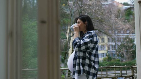 Téléchargez les photos : Contemplative femme enceinte se tient près du balcon appartement sirotant du thé en regardant la vue de la résidence, profitant d'un moment calme au cours du troisième trimestre - en image libre de droit