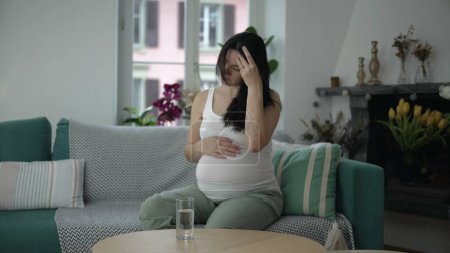 Foto de Mujer embarazada que experimenta molestias, señora de 30 años con náuseas del tercer trimestre sentada en el sofá del hogar - Imagen libre de derechos