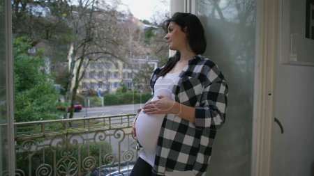 Foto de Mujer embarazada contemplativa acariciando tiernamente el vientre, de pie junto al balcón del apartamento con vistas a la vista con una sonrisa, encarnación del amor maternal - Imagen libre de derechos