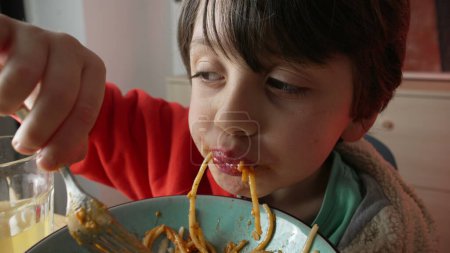 Child grabs fork and eats a mouthful of pasta spaghetti for supper, macro close-up face in wide angle, Italian food