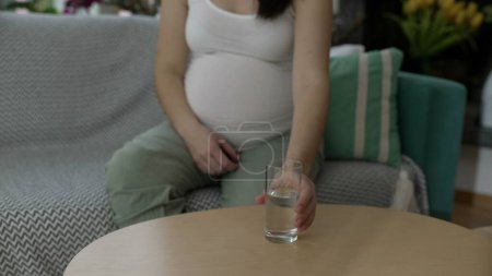 Photo for Pregnant woman picks up a glass of water seated on home couch, third trimester of pregnancy. Person taking care of health and wellbeing while expecting unborn baby - Royalty Free Image