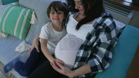 Tender child hugging pregnant mother in display of affection to unborn baby brother seated on couch at home