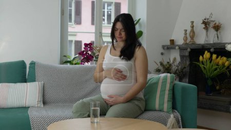 Foto de Mujer embarazada que tiene dolor, señora de 30 años luchando con náuseas durante el tercer trimestre de embarazo sentado en sofá sofá en la residencia en casa tocando suavemente el vientre - Imagen libre de derechos