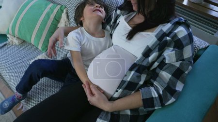 Foto de Abrazo cariñoso del niño a la madre embarazada, abrazando al hermano bebé no nacido en casa en el sofá - Imagen libre de derechos