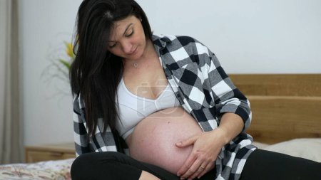 Foto de Mujer embarazada acariciando el vientre durante el tercer trimestre del embarazo sentada en la cama, etapa tardía - Imagen libre de derechos