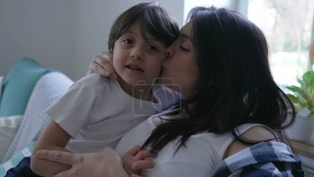 Photo for Mother and child healthy caring relationship in relaxing afternoon seated on couch at home, parent kissing son's cheek in display of love and affection - Royalty Free Image