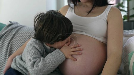 Heartwarming moment of 5 year old boy gently kissing mother's belly during late stage pregnancy, child awaiting baby unborn brother seated on couch at home with mom, maternal love concept