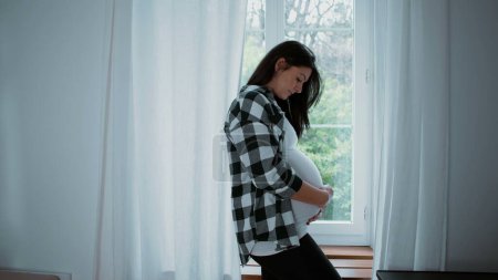 Glückseliger mütterlicher Moment, werdende Mutter streichelt 8-Monats-Bauch und erwartet Neugeborenes zu Hause am Fenster