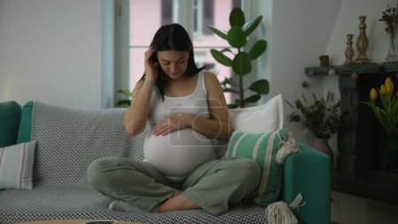 Pregnant 30s woman caressing belly seated on home couch sofa during third trimester pregnancy, maternal concept