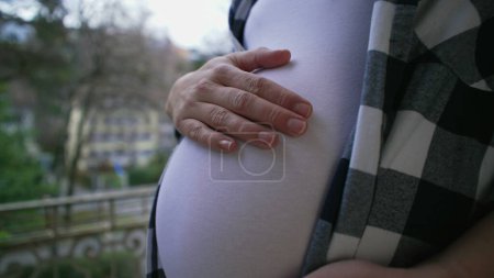 Foto de El amor maternal - la mujer embarazada que espera al bebé recién nacido acaricia el vientre de 8 meses por balcón del apartamento, caricia tierna de la mano en primer plano - Imagen libre de derechos