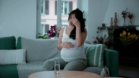 Foto de Mujer embarazada de 30 años lucha con dolor de cabeza y dolor sentado en casa sofá sofá residencia, concepto de salud materna - Imagen libre de derechos