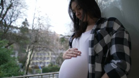 Téléchargez les photos : Mère enceinte caressant 8 mois ventre par appartement balcon montrant l'amour et l'affection attendant le nouveau-né - en image libre de droit