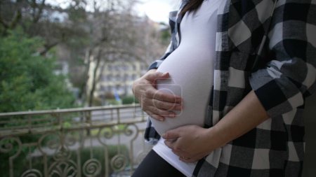 Téléchargez les photos : Mère enceinte caressant 8 mois ventre par appartement balcon montrant l'amour et l'affection attendant le nouveau-né - en image libre de droit