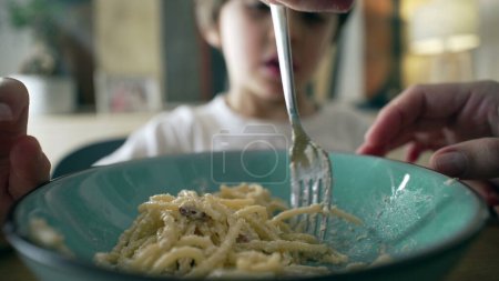 Close-up of fork spinning spaghetti on blue plate with little boy in blurred background, mother's hand teaching son to spin pasta, child's dish mealtime