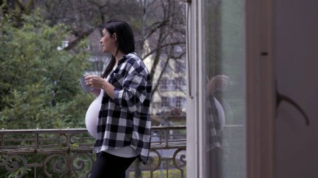 Foto de Escena pacífica de una mujer embarazada bebiendo té de pie junto al balcón del apartamento mirando a la vista en contemplación, el tercer trimestre de embarazo esperando al bebé recién nacido - Imagen libre de derechos