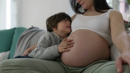 Foto de Escena familiar conmovedora: la madre embarazada acaricia tiernamente a su hijo de 5 años en el sofá, capturando un momento de afecto en el embarazo tardío - Imagen libre de derechos