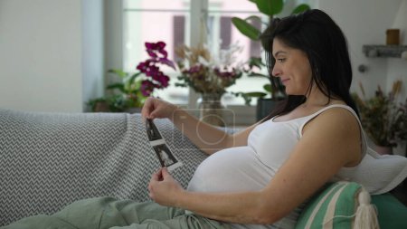 Happy Pregnant 30s woman seated on couch looking at ultrasound pictures of unborn baby, expecting child in third trimester pregnancy