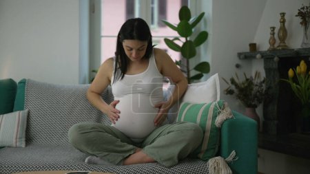 Foto de Mujer embarazada de 30 años acariciando vientre sentado en el sofá del sofá del hogar durante el embarazo del tercer trimestre, concepto materno - Imagen libre de derechos