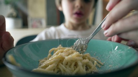 Close-up of fork spinning spaghetti on blue plate with little boy in blurred background, mother's hand teaching son to spin pasta, child's dish mealtime