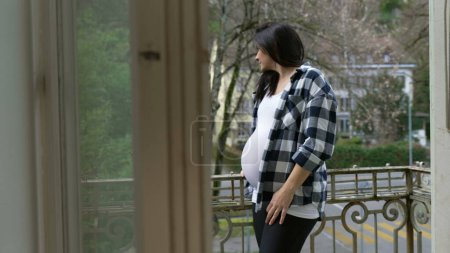 Téléchargez les photos : Pensive 30s femme enceinte se tient au balcon de l'appartement en ajustant les cheveux et en regardant la vue de la maison de résidence dans l'introspection calme - en image libre de droit