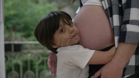 Téléchargez les photos : Mignon petit garçon étreignant ventre enceinte de la mère montrant l'amour et l'affection pendant le stade tardif de la grossesse - en image libre de droit