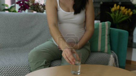 Téléchargez les photos : Femme enceinte Hydratante, ramasse un verre d'eau, Assise sur le canapé avec du verre d'eau dans le troisième Trimester prenant soin de la santé et du bien-être - en image libre de droit
