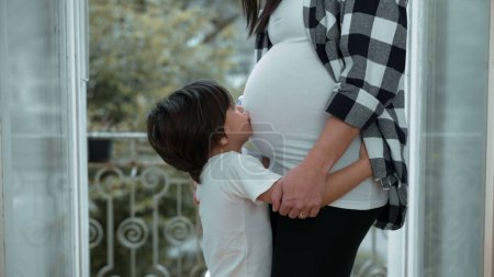 Niño pequeño abrazando el vientre de la madre embarazada por el balcón, afecto tierno por el hermano nonato