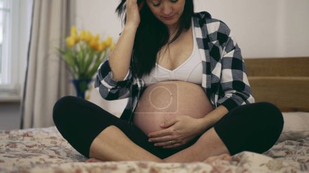Téléchargez les photos : Troisième trimestre grossesse - femme caresse ventre enceinte en 8 mois s'attendant bébé assis dans le lit toucher doucement ventre - en image libre de droit