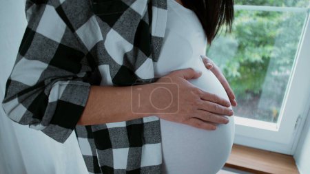 Foto de Hermoso momento materno de madre embarazada acariciando su vientre de 8 meses esperando con anticipación a su recién nacido de pie junto a la ventana - Imagen libre de derechos