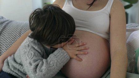 Herzerwärmender Moment des 5-jährigen Jungen, der den Bauch der Mutter während der späten Schwangerschaft sanft küsst, Kind erwartet ungeborenen Bruder, der zu Hause mit Mama auf der Couch sitzt, mütterliches Liebeskonzept