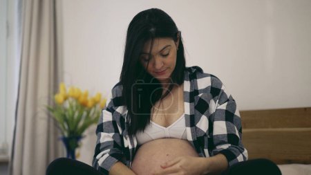 Foto de Tercer trimestre de embarazo - mujer acaricia vientre embarazada en 8 meses esperando bebé sentado en la cama suavemente tocando el vientre - Imagen libre de derechos