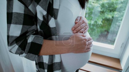 Foto de Hermoso momento materno de madre embarazada acariciando su vientre de 8 meses esperando con anticipación a su recién nacido de pie junto a la ventana - Imagen libre de derechos