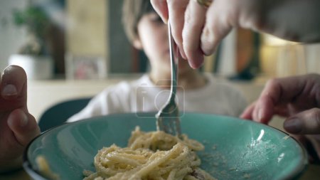 Großaufnahme der Gabel, die Spaghetti auf blauem Teller spinnt, mit kleinem Jungen im verschwommenen Hintergrund, die Hand der Mutter bringt dem Sohn bei, Nudeln zu spinnen, die Mahlzeit des Kindes