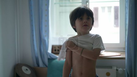 Téléchargez les photos : Enfant mettant t-shirt, jeune garçon habiller par lui-même debout dans les vêtements de chambre à coucher chemise blanche. 5 ans caucasien mâle enfant - en image libre de droit