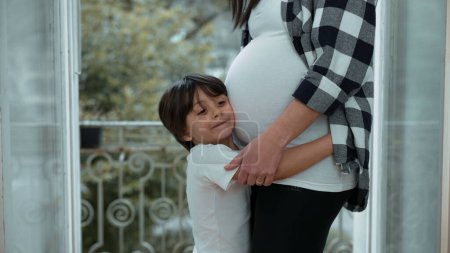 Kleiner Junge umarmt den Bauch der werdenden Mutter vom Balkon, zarte Zuneigung für ungeborene Geschwister